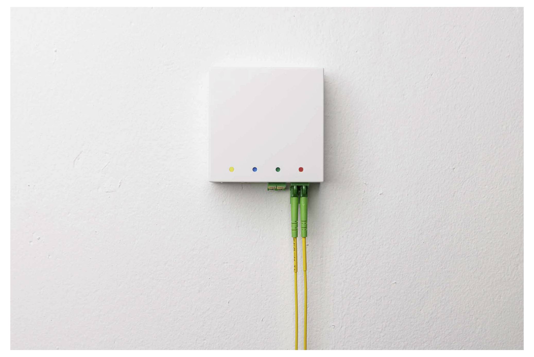 Hvidt vægmonteret fiberoptisk netværkskab med to gule kabler og fire farvede statusindikatorer nedenunder.