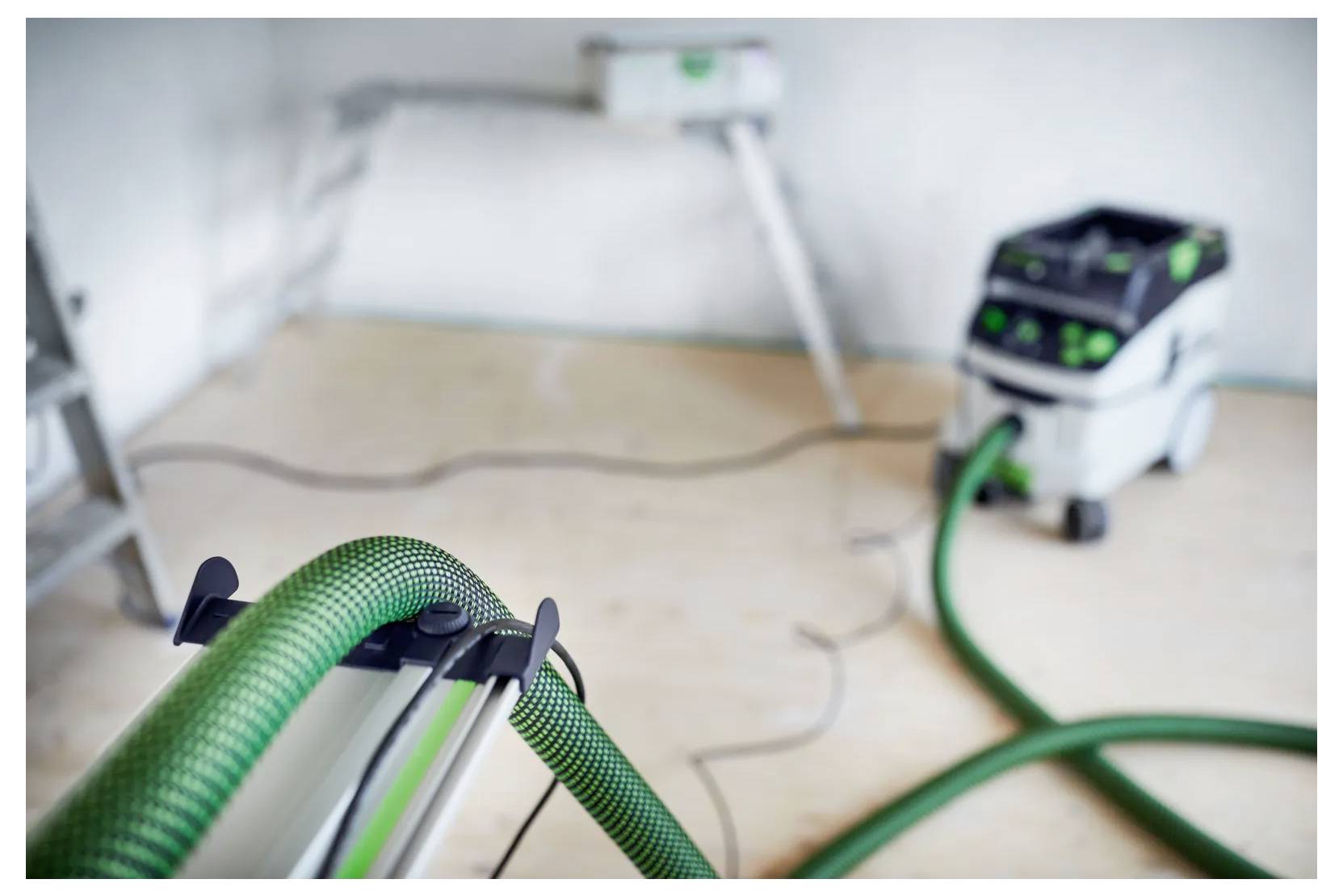 Vakuumsliber med grønt slange i et værelse under renovering, med slibeudstyret i forgrunden og en støvudsuger i baggrunden.