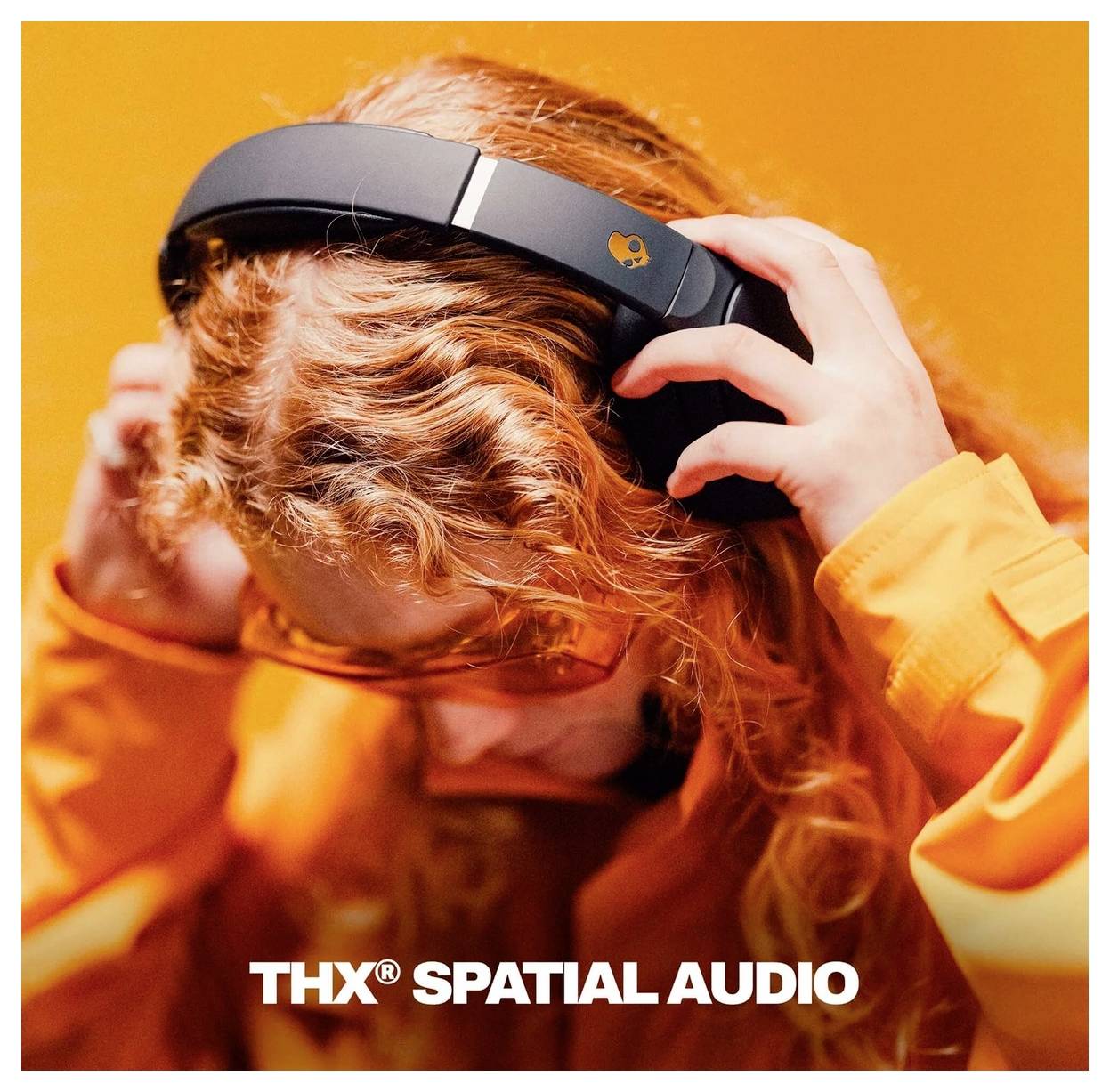 Person med krøllet rødt hår, der bærer hovedtelefoner mod en orange baggrund, med teksten 'THX SPATIAL AUDIO', som indikerer immersive lyd.