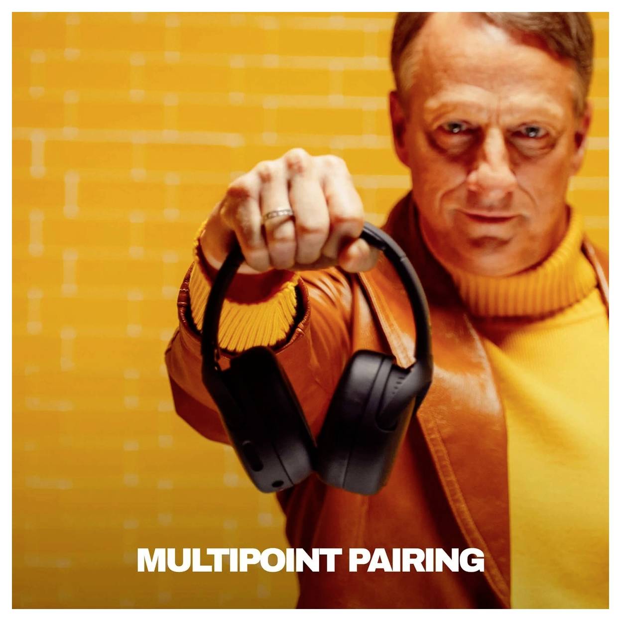 Mand i orange jakke holder sorte trådløse hovedtelefoner mod kameraet, med teksten "MULTIPOINT PAIRING" nederst.