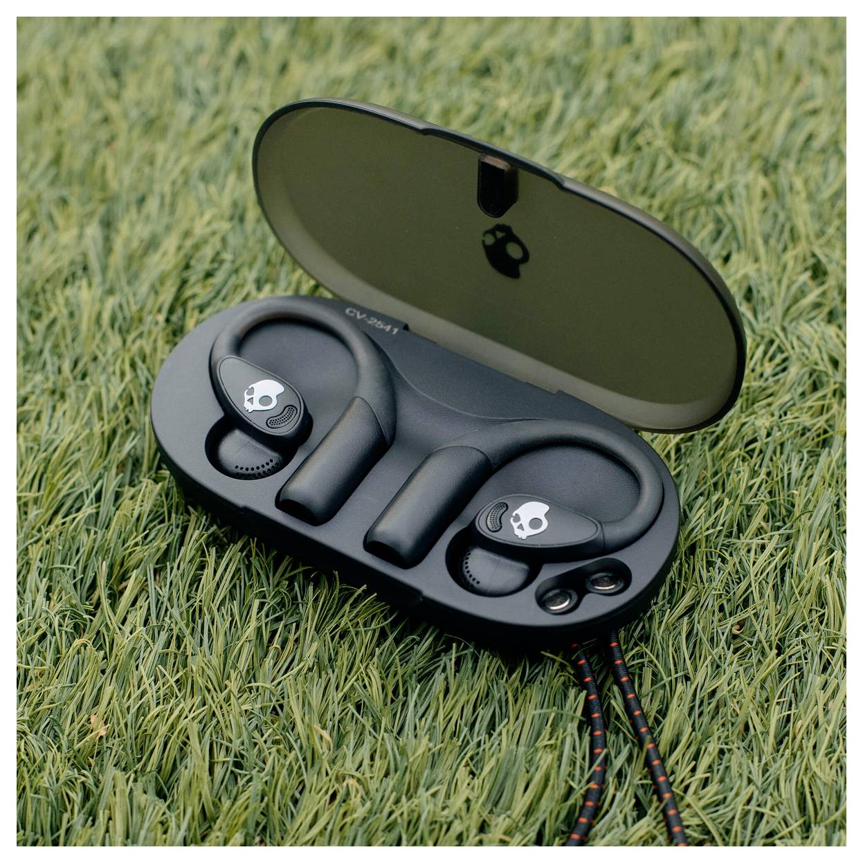 Åben etui med sorte Skullcandy trådløse øretelefoner på græs med opladningskabel tilsluttet.