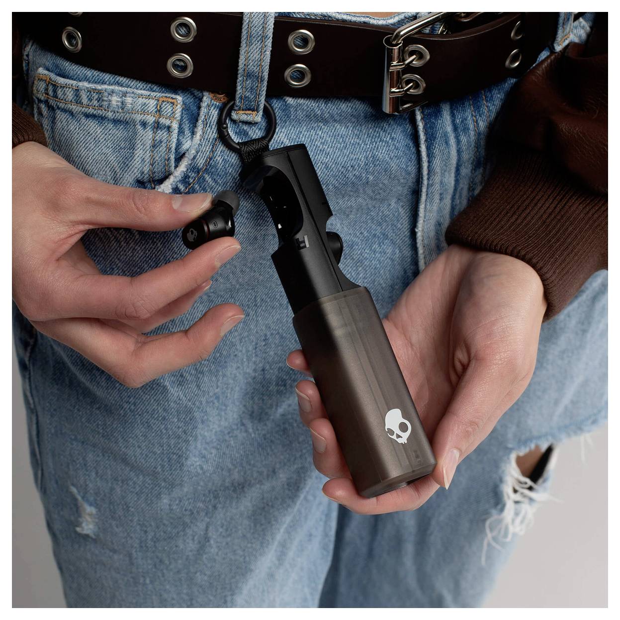 Tætbillede af en person, der holder en håndholdt enhed med et dødningelogo og indsætter en øretelefon, iført jeans og bælte.