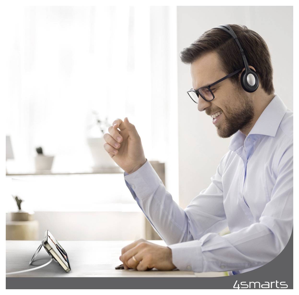 Smilende mand med briller og headset løfter sin knyttet hånd under et videoopkald, siddende ved et skrivebord med en smartphone på en holder.
