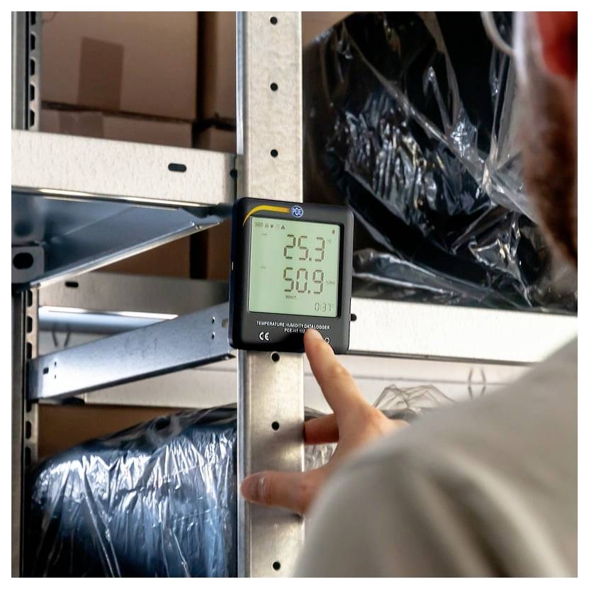 En person tjekker et digitalt termometer på et metallageringsreol; displayet viser 25,3 og 50,9, sandsynligvis temperatur og luftfugtighed.