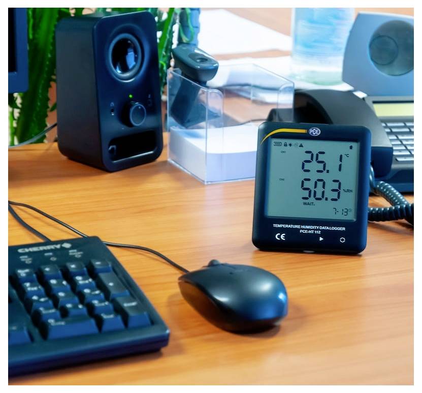 En digitalt temperatur- og fugtighedsmåler på et skrivebord viser 25,1°C og 50,3% fugtighed, ved siden af et tastatur, en mus og en højttaler.