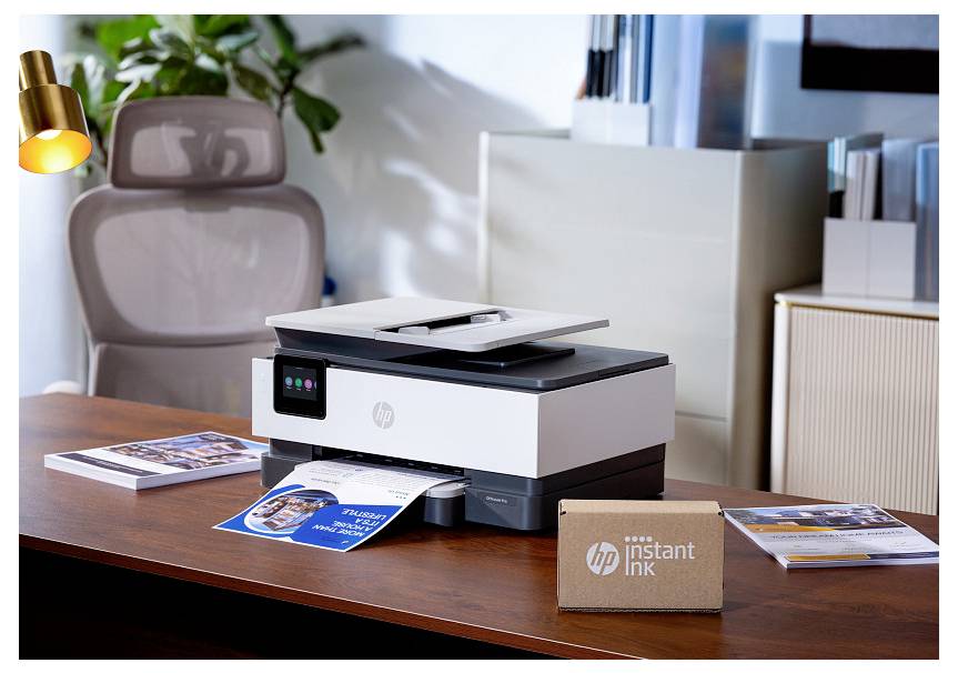 HP multifunktionsprinter, der udskriver farvefoldere på et skrivebord, med en Instant Ink-kasse ved siden af i et hjemmekontorsetup.
