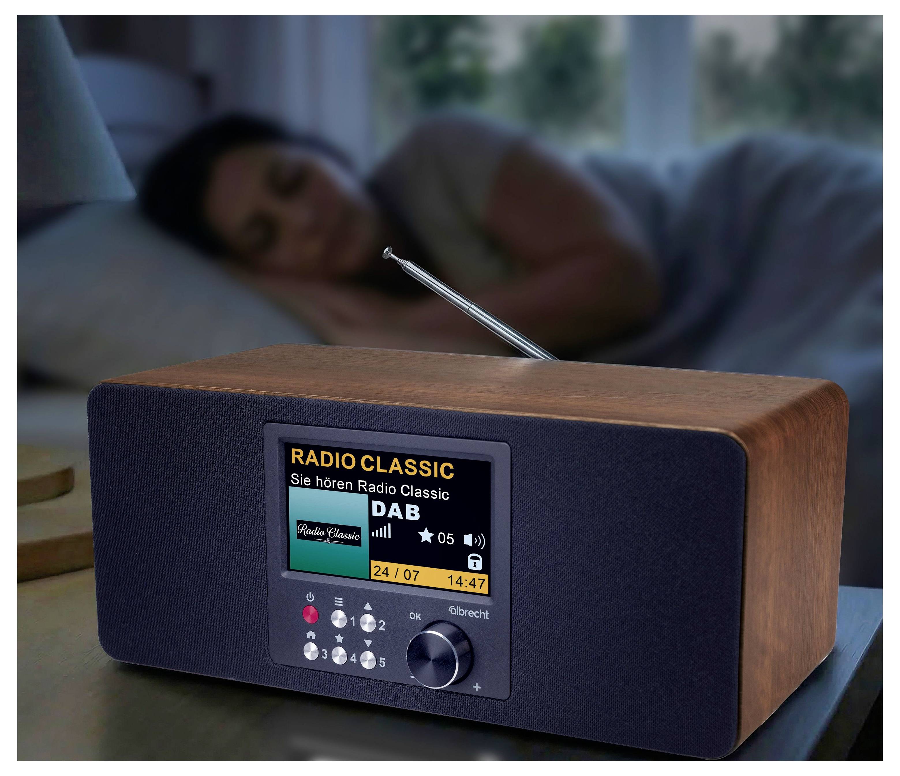 Digitalt radio på sengebord, der afspiller 'RADIO CLASSIC' DAB, med en sovende person uskarpt i baggrunden.