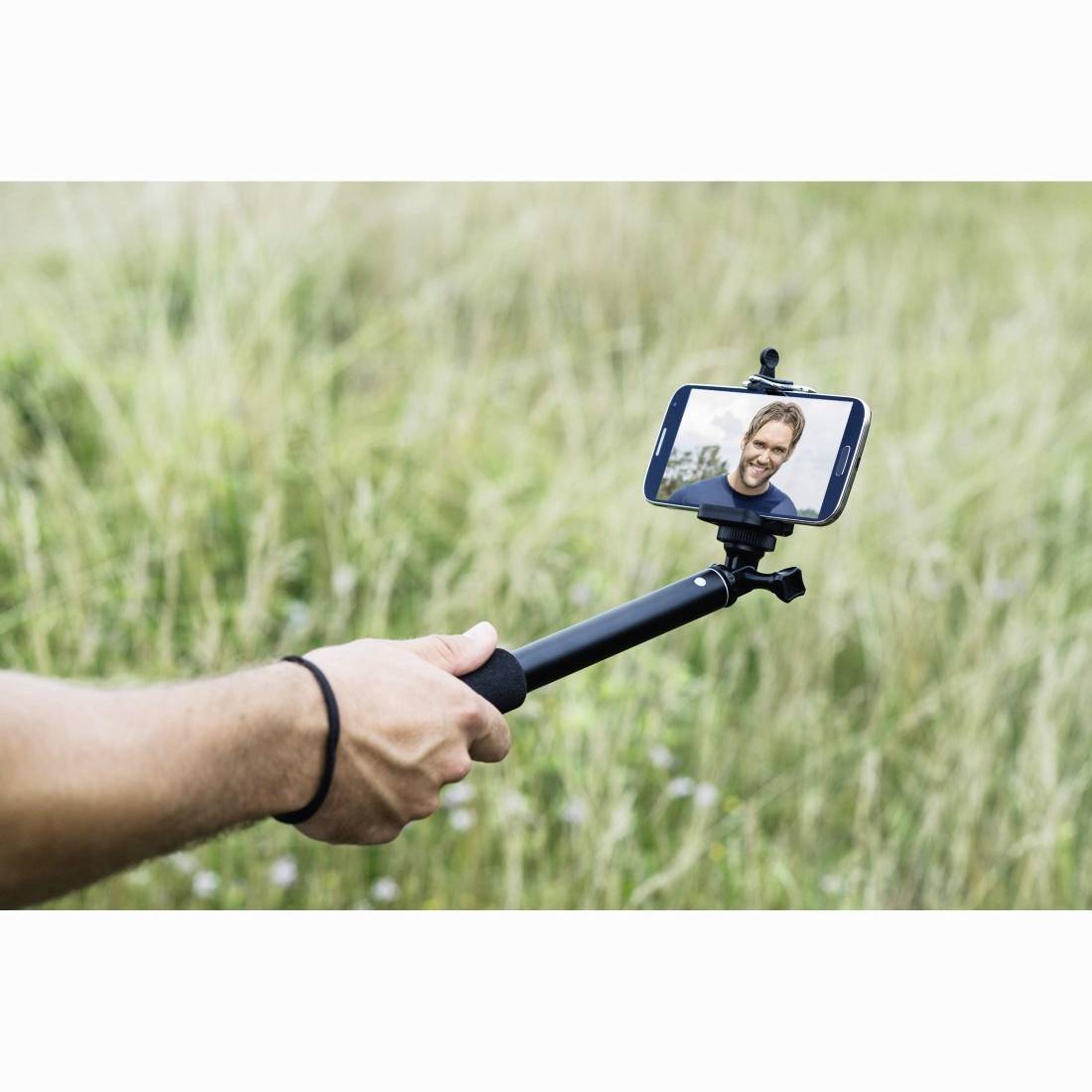 En person, der holder en selfie-stok med en smartphone, der viser en smilende mand på skærmen, udendørs i højt græs.
