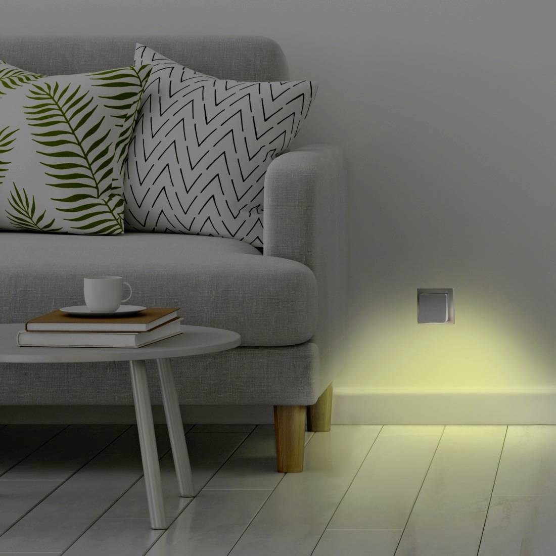 Moderne stue med grå sofa, mønstrede puder, sidetabel med bøger og kop, og en væglampe, der kaster et blødt skær på gulvet.