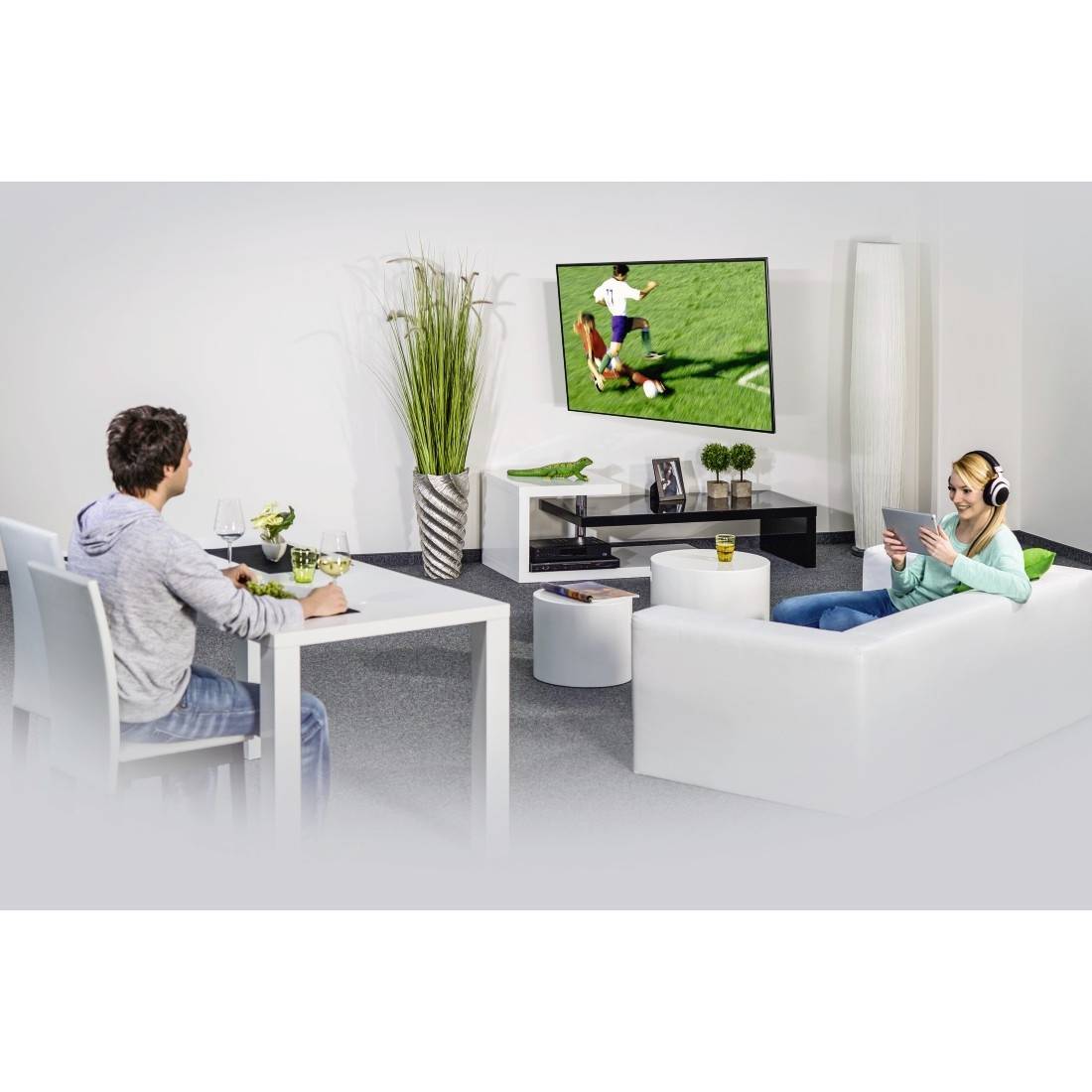 To mennesker hviler sig i en stue og ser en fodboldkamp på et tv monteret på væggen, den ene sidder ved et spisebord og den anden ligger på en sofa med hovedtelefoner.