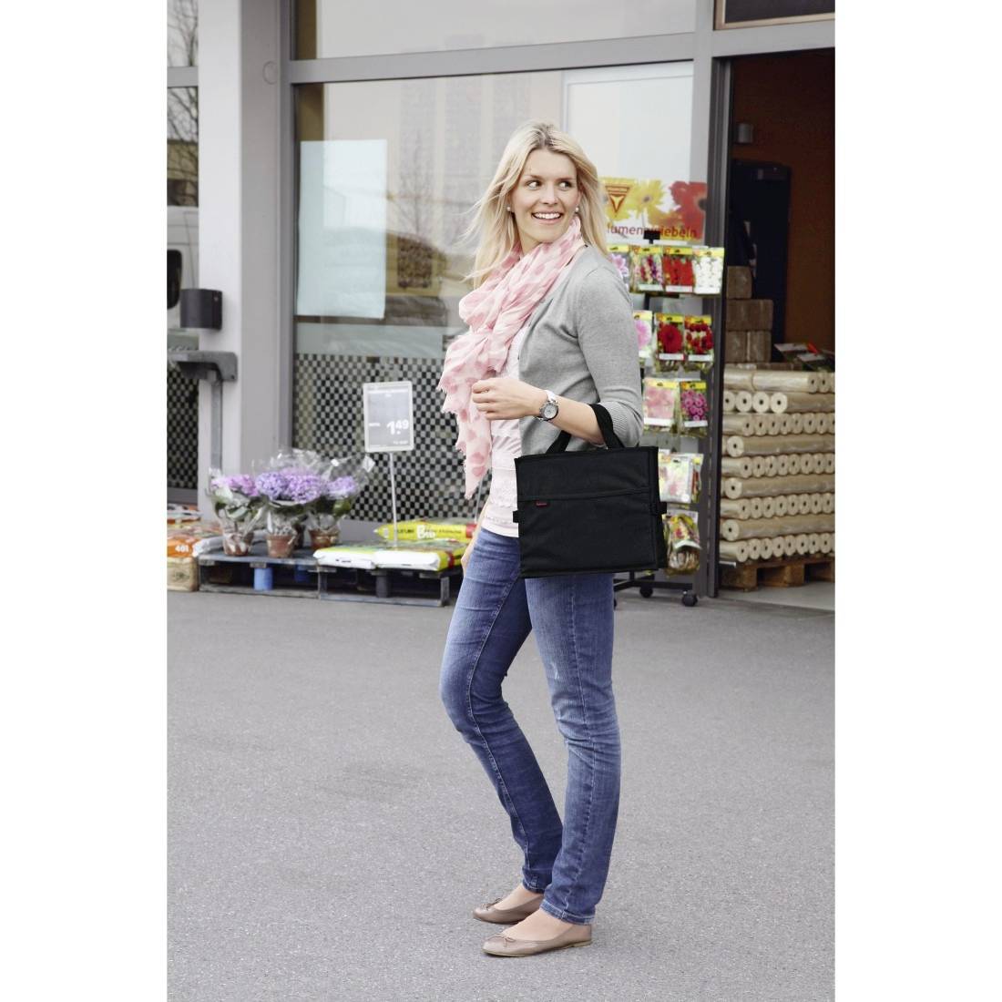 Blond kvinde i en grå sweater, pink tørklæde og jeans står uden for en butik og holder en sort tote-taske.