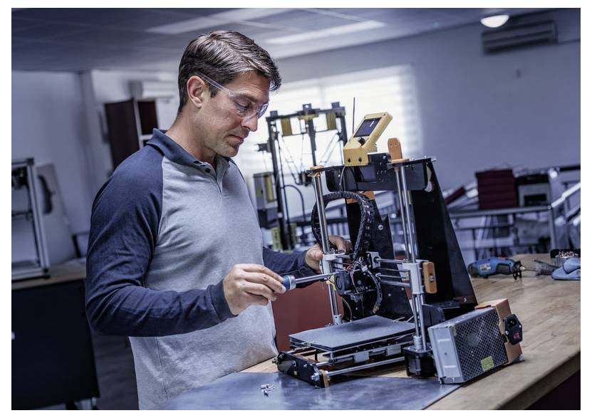 En mand justerer en 3D-printer på en arbejdsbænk i et værksted og bruger et værktøj til at reparere eller kalibrere maskinen.