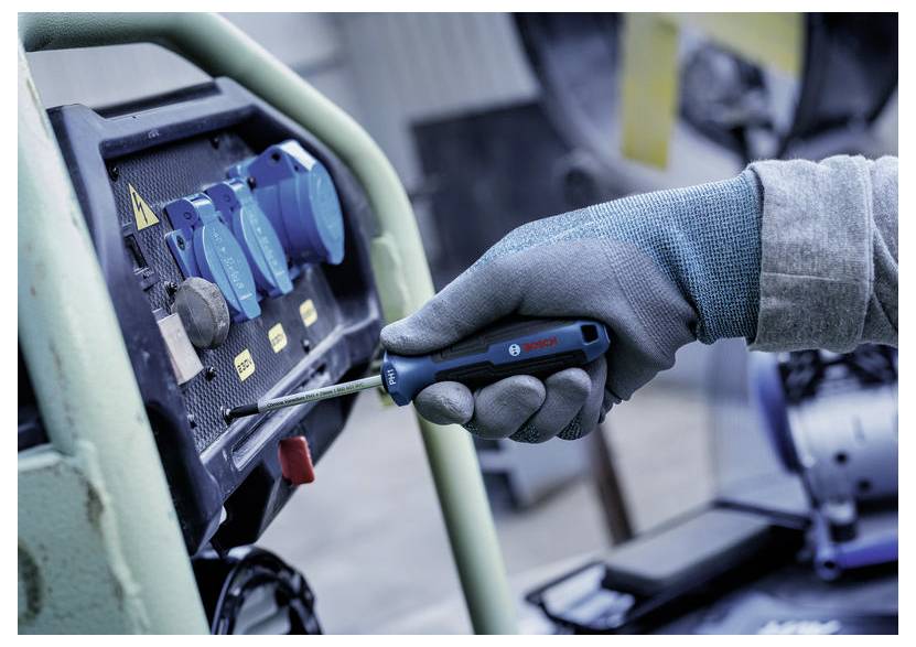Hånd med handsker, der bruger en skruetrækker på et elektrisk kontrolpanel med blå kontakter og en rød knap, hvilket tyder på vedligeholdelse eller reparation af udstyr.