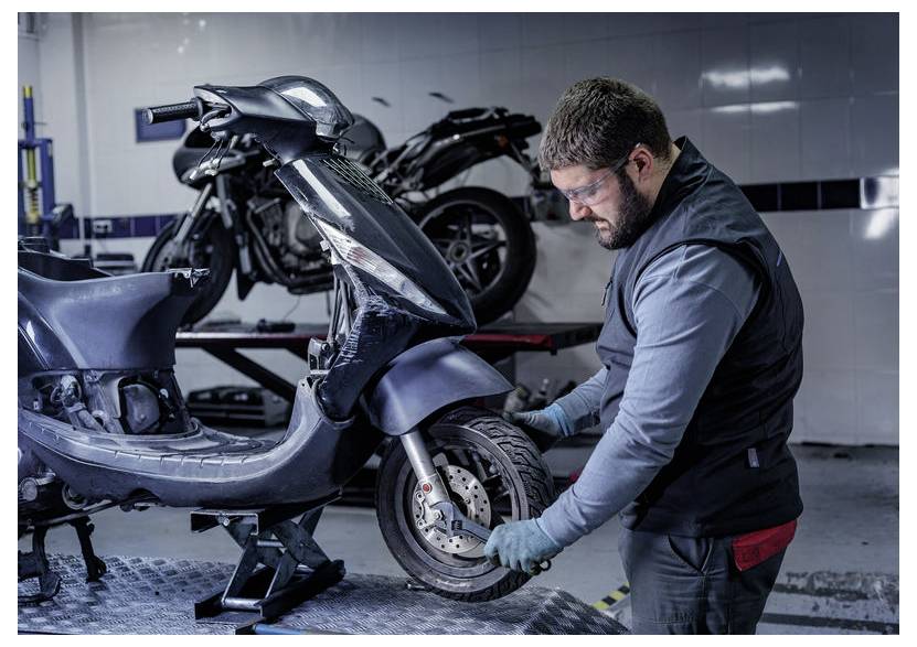 En mekaniker i et værksted reparerer forhjulet på en sort scooter på en lift, med en anden motorcykel i baggrunden.