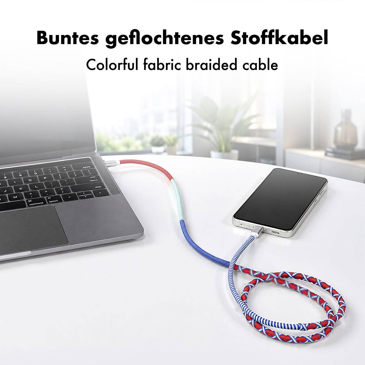 Farverigt flettet opladningskabel, der forbinder en bærbar computer og smartphone på et skrivebord, med teksten "Buntes geflochtenes Stoffkabel" og "Colorful fabric braided cable".