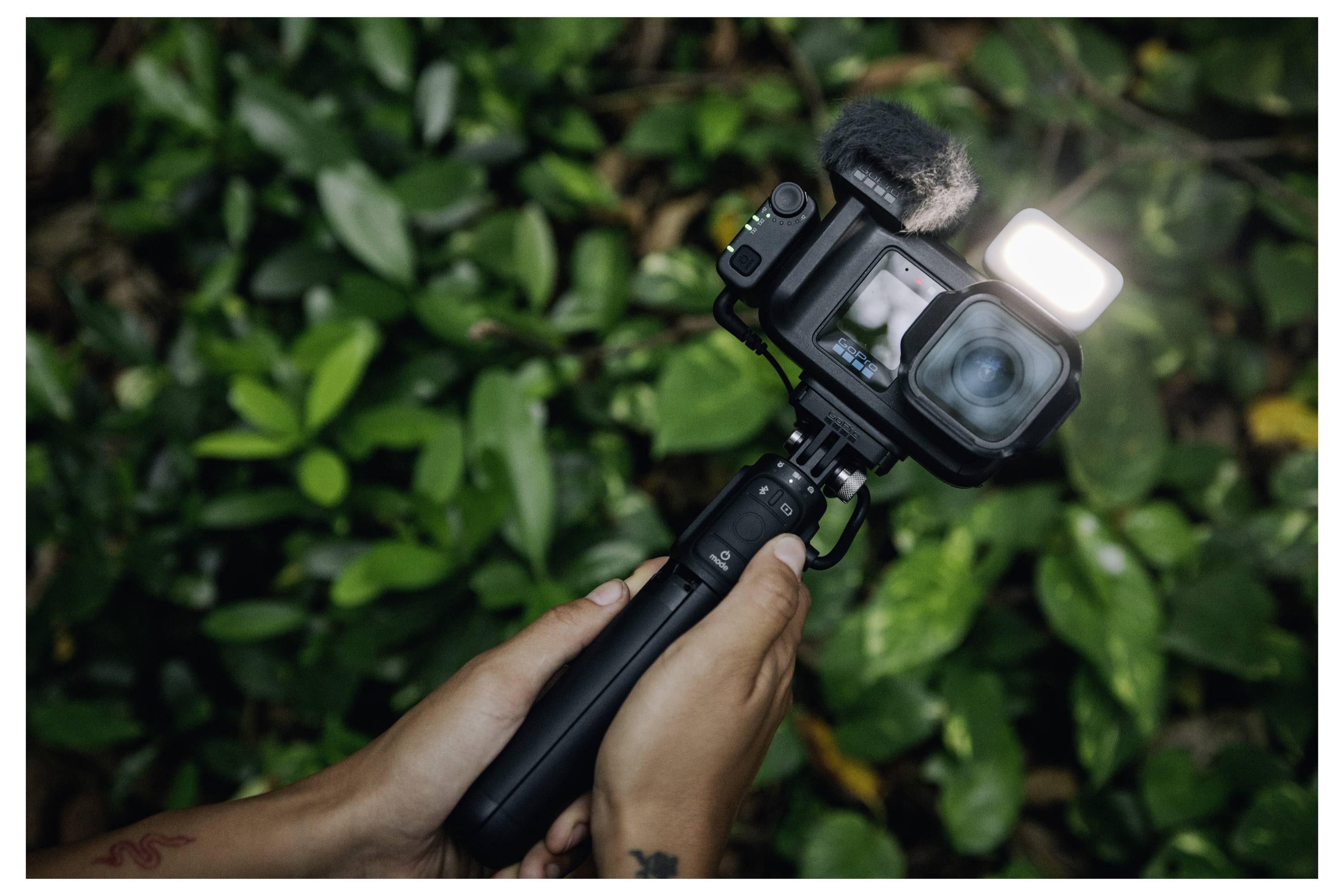Hænder, der holder et vlogging-actionkamera på en håndholdt greb, med dets lys tændt, rettet mod bladrig grønne plantedele.