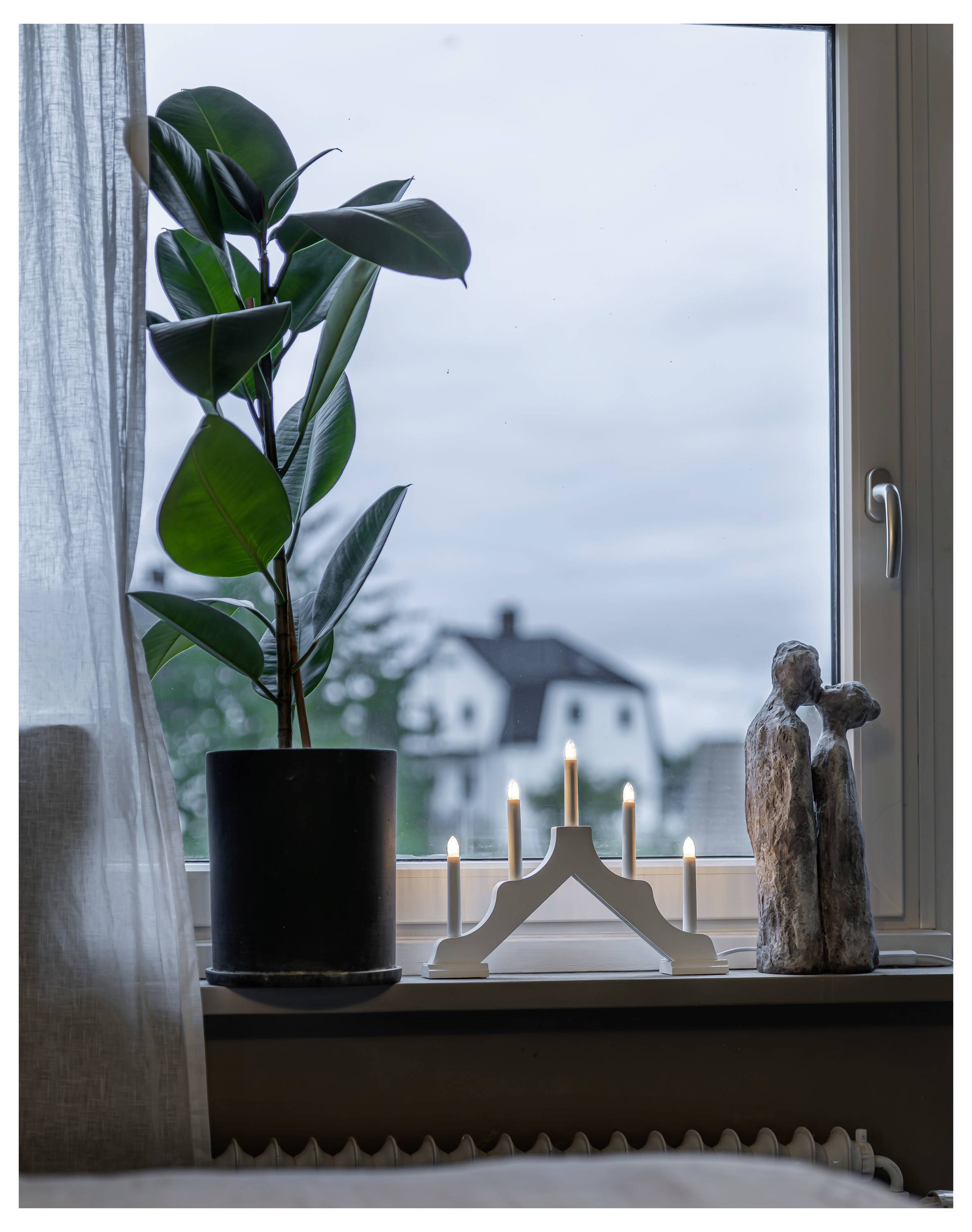 Plante i potte og tændte stearinlys på en vindueskarm ved siden af en lille træskulptur, med et hus synligt udenfor vinduet.