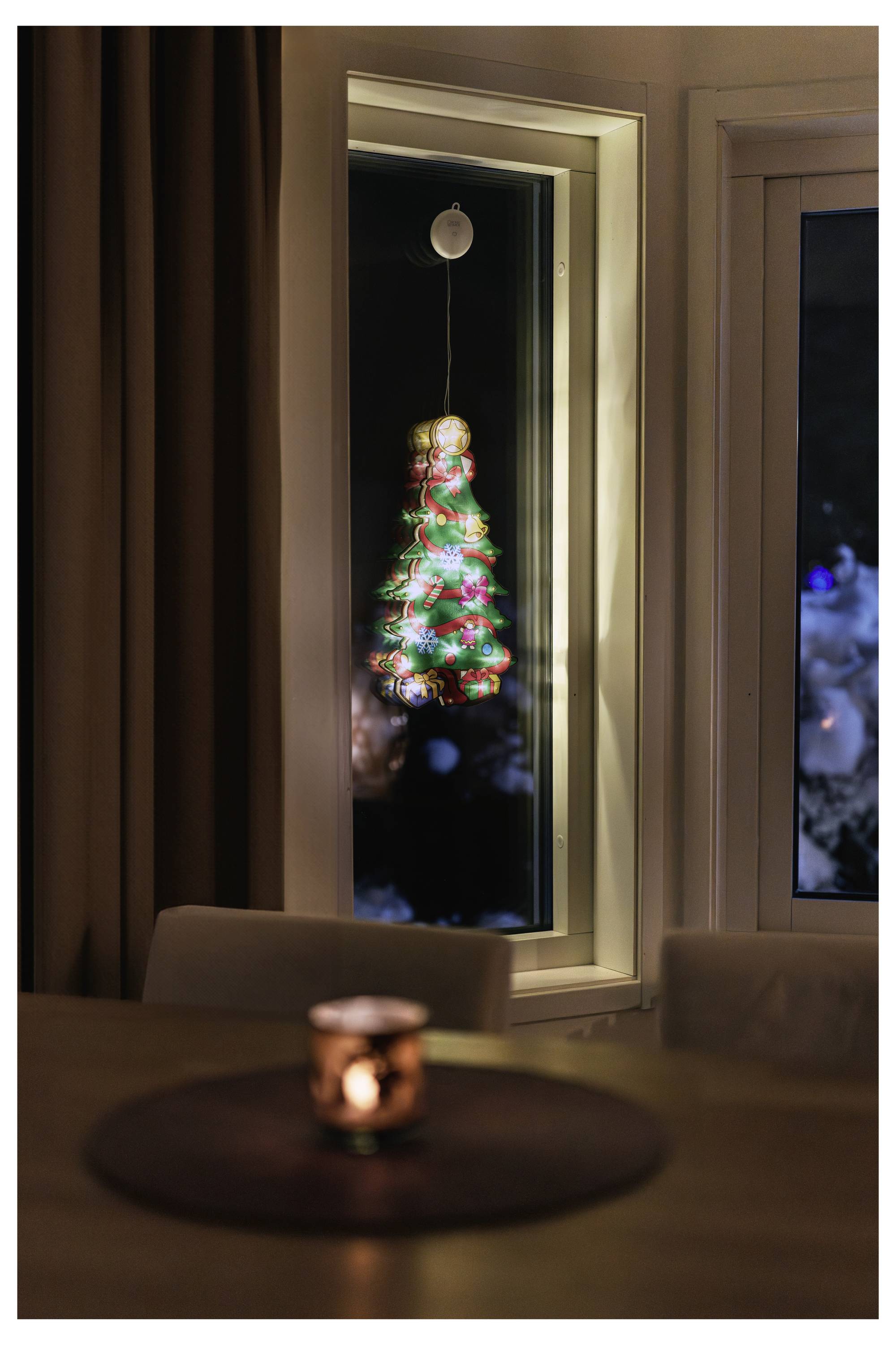 En lysende juleudsm6kning hænger uden for et vindue om natten, reflekteret i glasset over et stearinlysbeklædt bord indendørs.