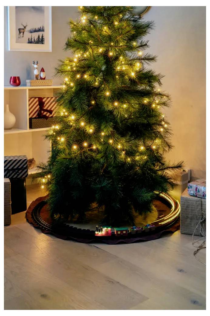 Et dekoreret indendørs juletre med varme hvide lys står på et træskjul i et hyggelig værelse med hylder og indpakkede gaver i nærheden.