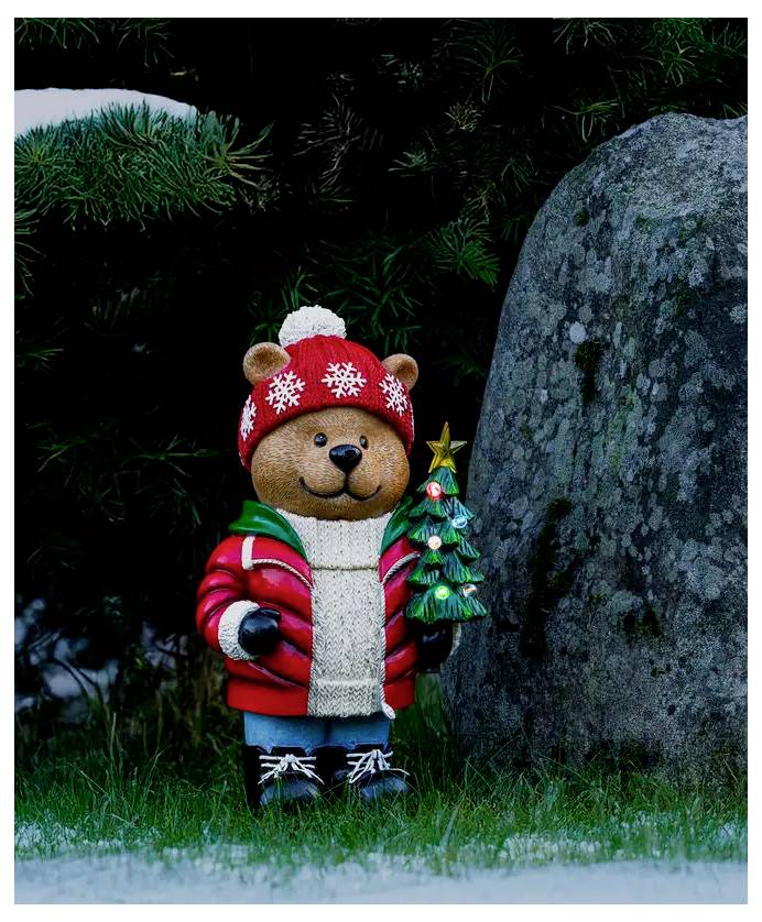 En teddybjørne-figur i en rød vinterkasket og frakke holder et lille tændt juletræ ved siden af en stor sten udendørs.
