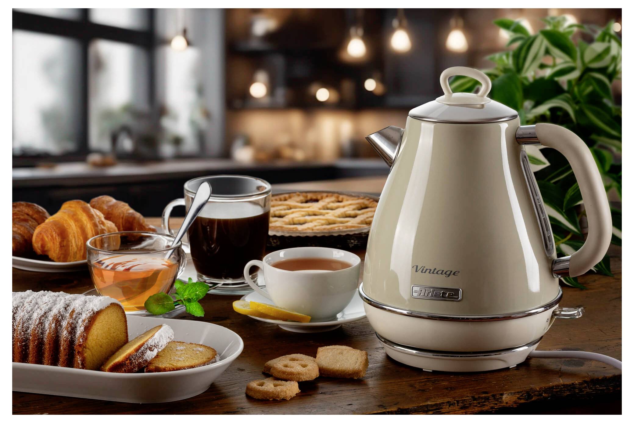 Køkkenbord med vintage elektrisk kedel, kaffekrus, te, croissanter, kage, småkager og en pottet plante i baggrunden.