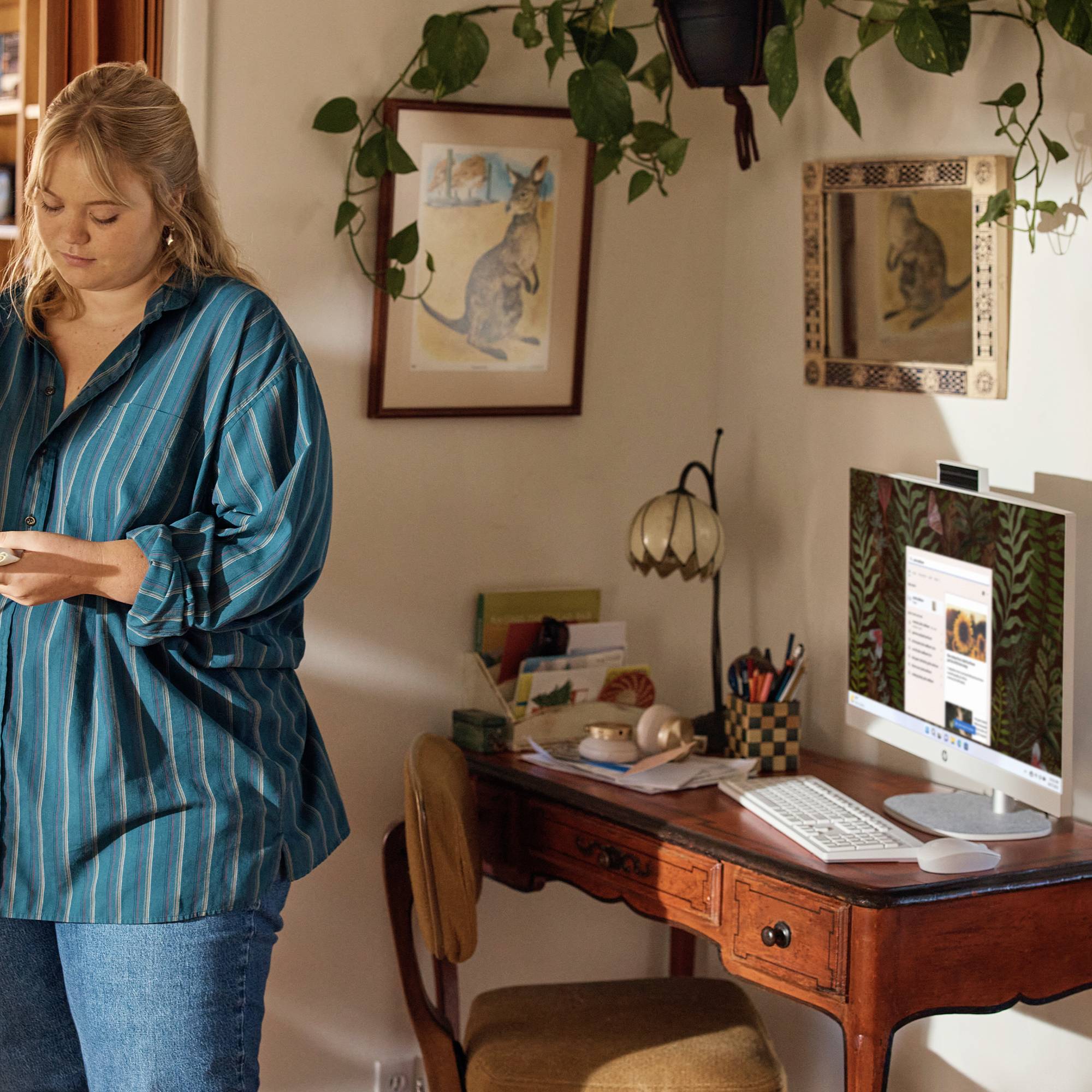 Kvinde, der står i et hjemmekontor, kigger på en telefon ved siden af et skrivebord med en iMac, papirer og hængende planter.