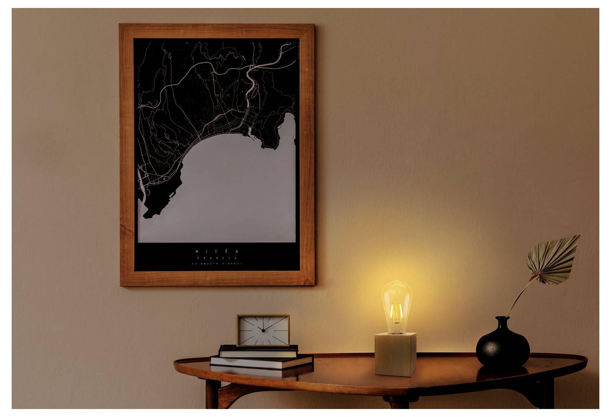 Et indrammet kort hænger over et lille bord med en glødende lampe, bøger, ur og vase, hvilket skaber en varm interiørscene.