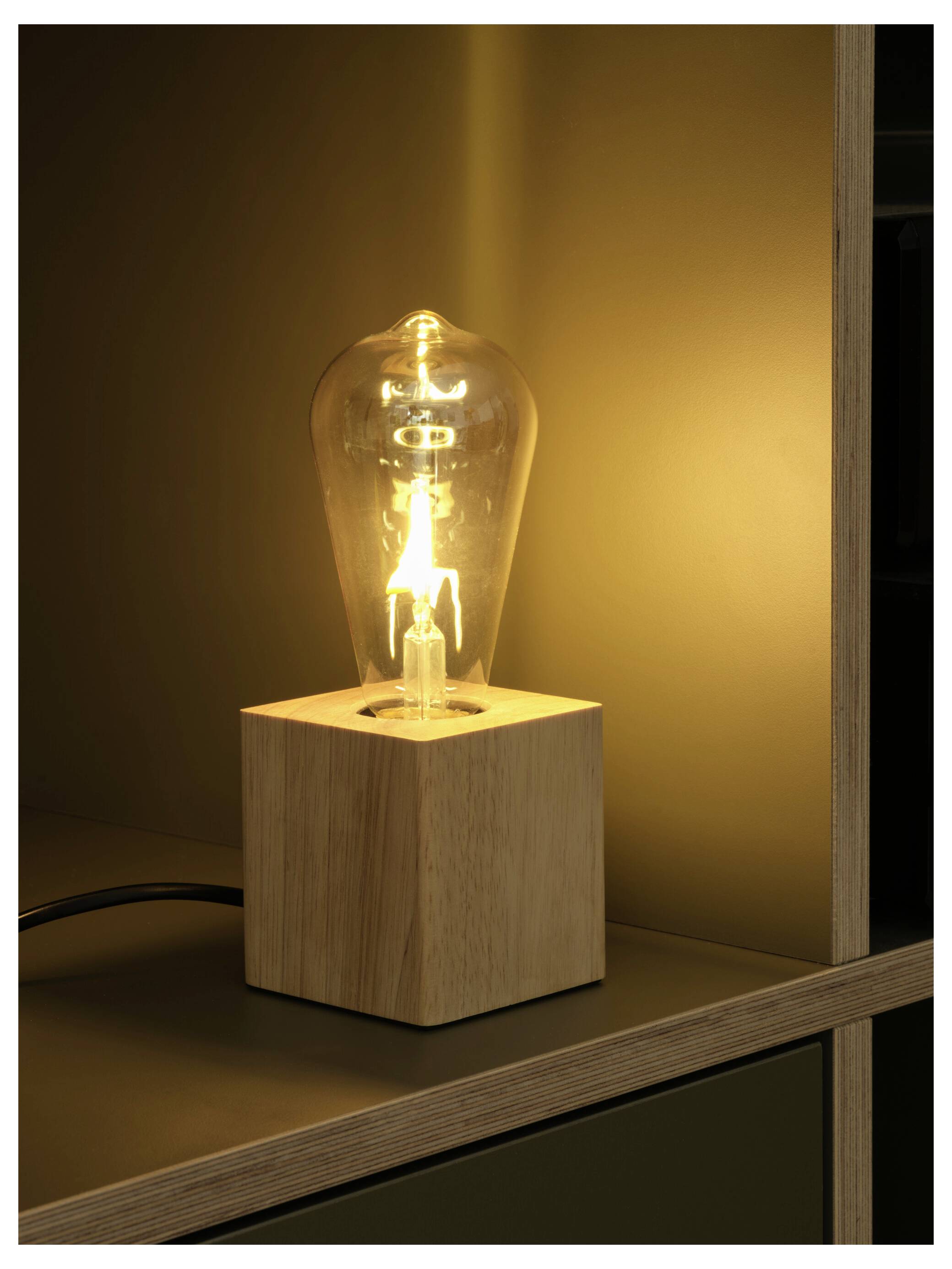 Varm glødende Edison-stil pæreligtlampe i en træ-kubisk sokkel på en hylde mod en mørk væg.
