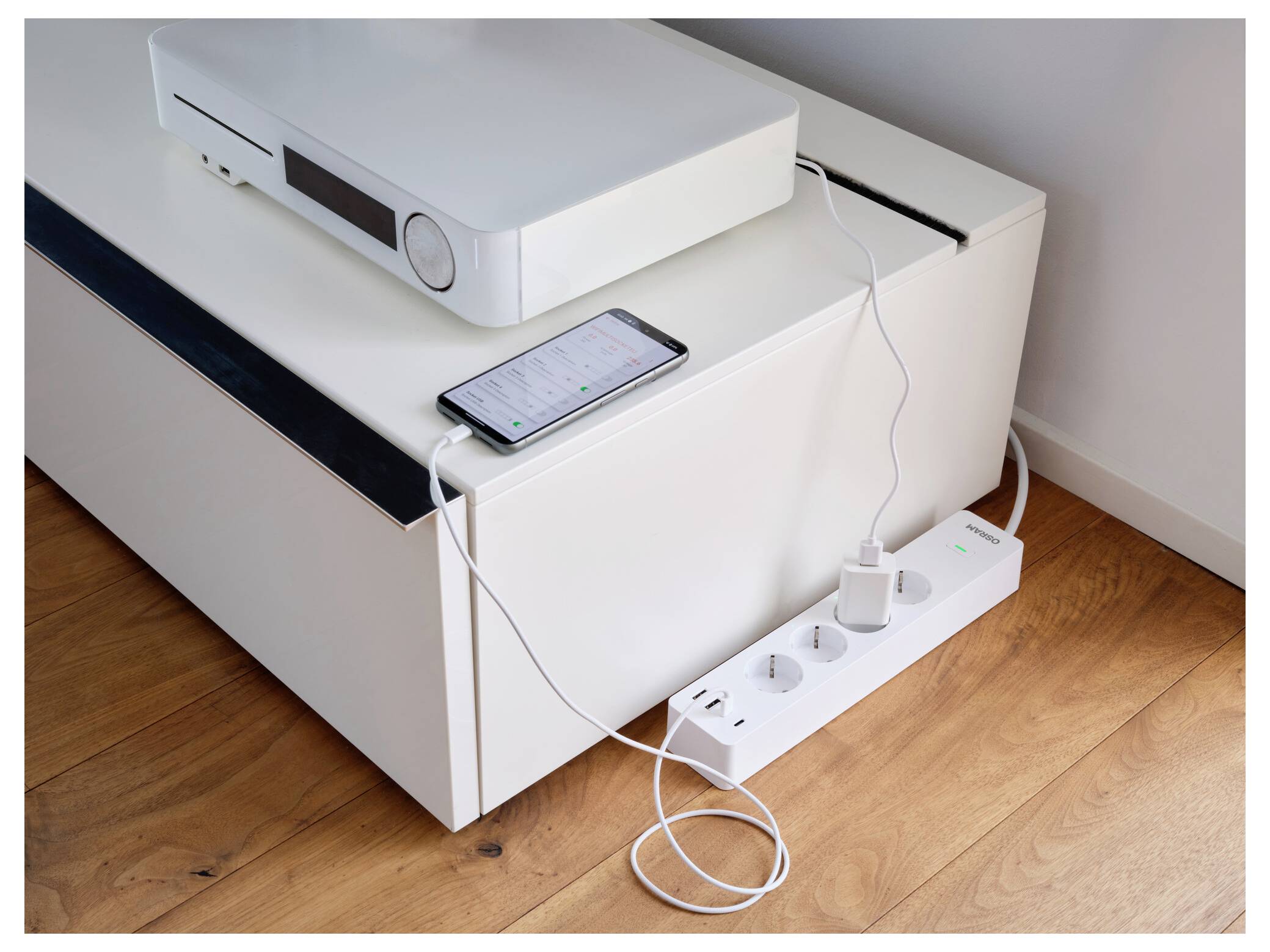 Hvid projektor på et skab med en smartphone, der lader ved siden af det, og en strimmelkontakt nedenfor, der viser forbundne kabler i en hjemmeopstilling.