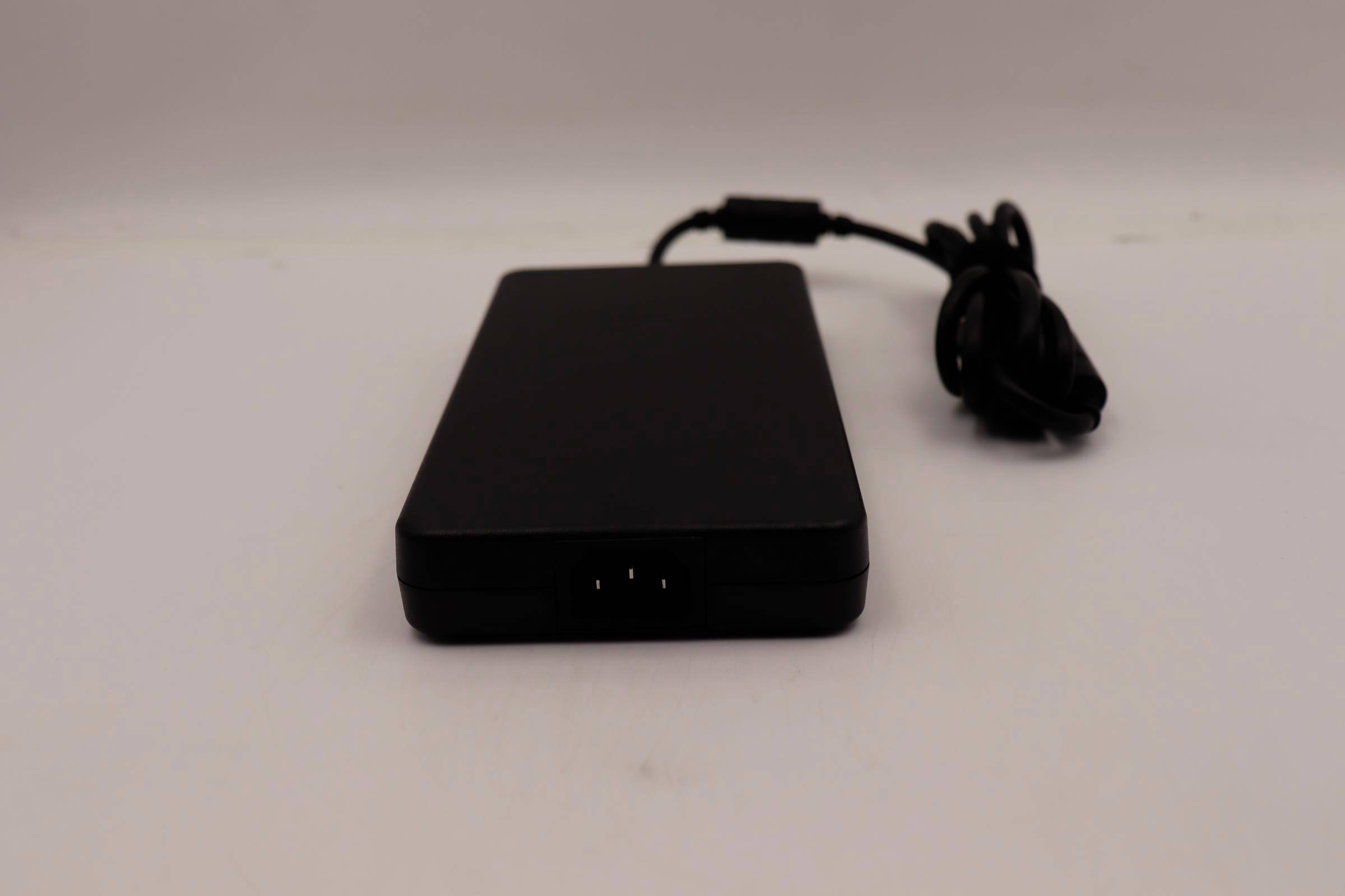 Sort rektangulært elektronisk apparat eller ekstern harddisk med tilsluttet kabel, hvilende på en hvid overflade.