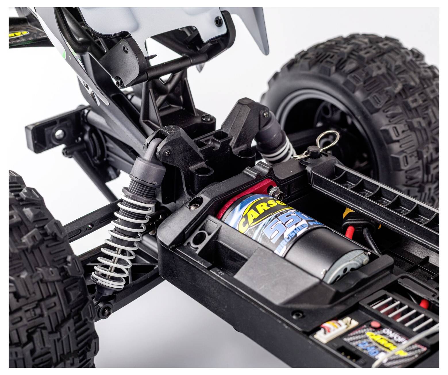 Tætbillede af et off-road RC-bilchassis med ophæng, motor og store knobbede dæk på en hvid baggrund.