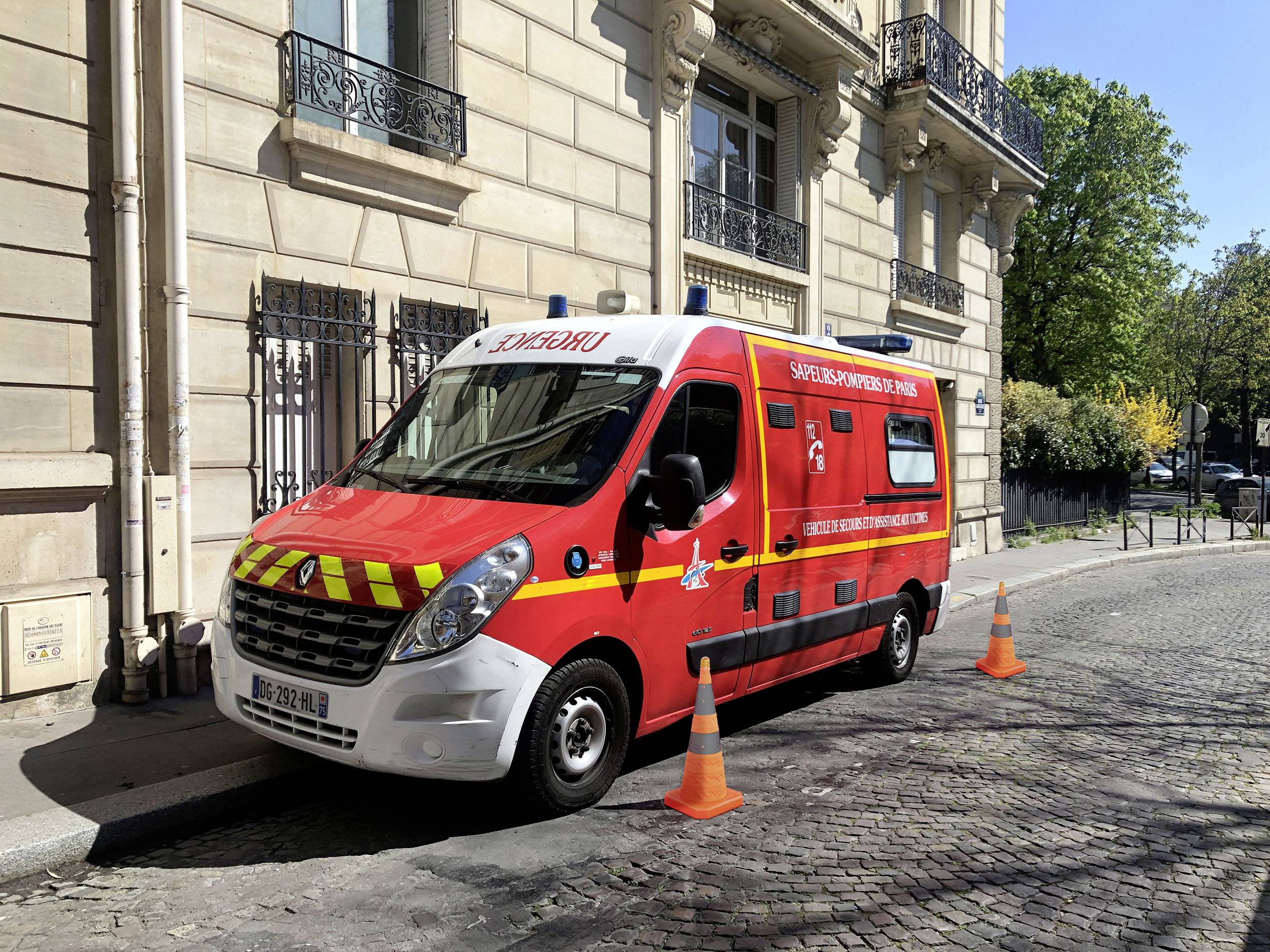 Rød beredskabsvogn parkeret på en brosten gade ved siden af en bygning, mærket 'SAPEURS-POMPIERS' med trafikkeglers i nærheden.