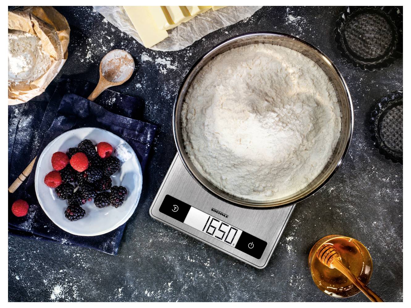 Køkkenschene med mel i en røreskål på en digital vægt, der viser 150, omgivet af bær, køkkenredskaber og bageingredienser på en melet overflade.