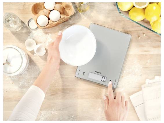En hånd placerer en hvid skål på en digital køkkenvægt omgiver af bagingredienser, med æg, mel, mælk og citroner på en bordplade.