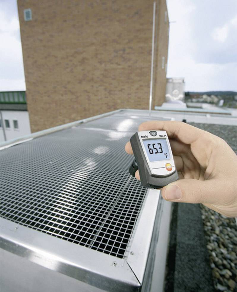 En hånd holder et digitalt termometer, der viser 63 grader Fahrenheit, over et udvendigt tagrist på en bygning. Baggrunden viser himmel og bygninger.