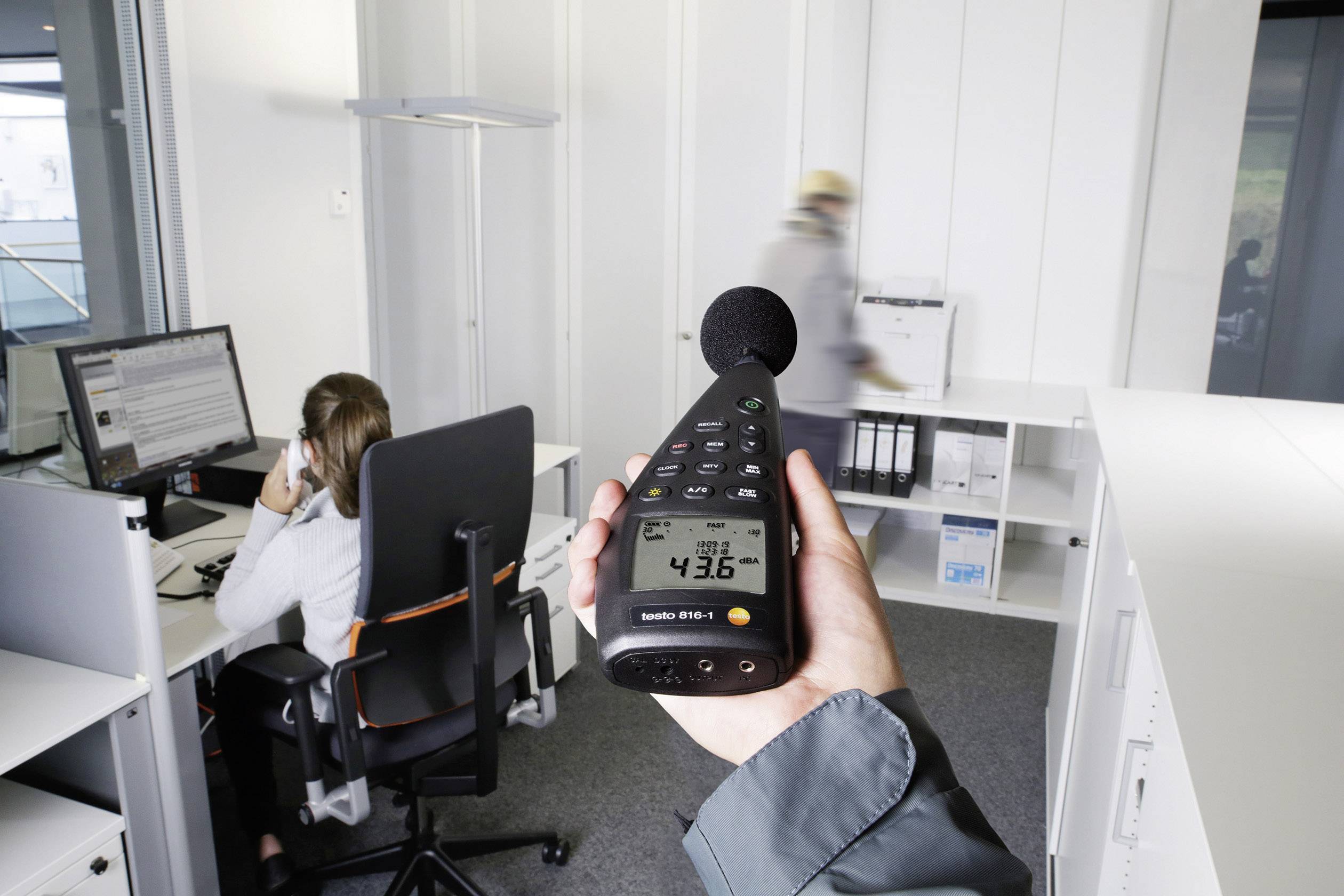 En person holder en decibelmåler i et kontor. Måleapparatet viser 43,6 decibel. I baggrunden arbejder personer ved skriveborde.