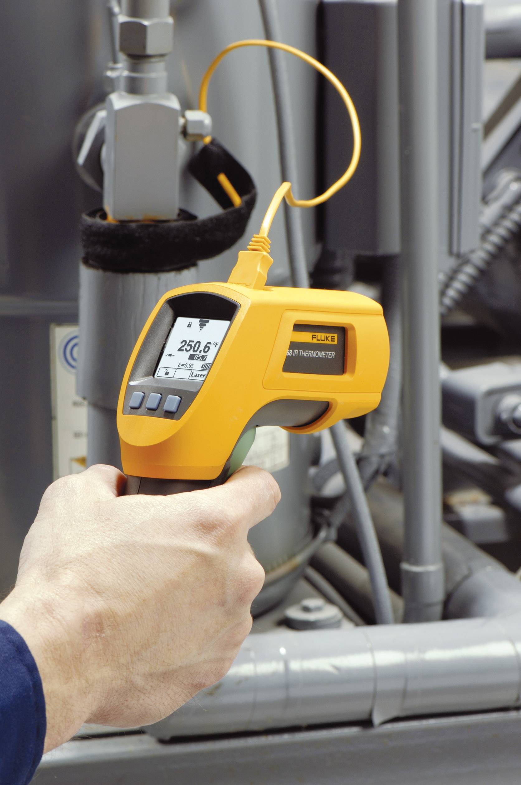 En hånd holder et infrarødt termometer, der måler en temperatur på 25,6 grader celsius foran en industriel maskine.