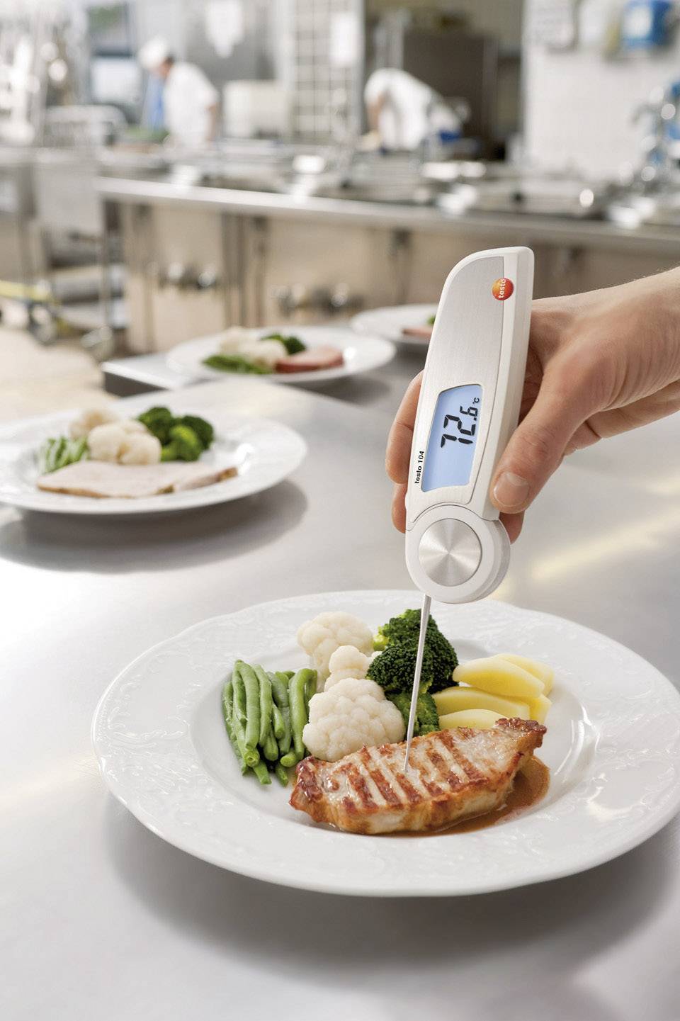 Et termometer måler temperaturen på et grillet kyllingefilet på en tallerken med grøntsager, formentlig i et restaurantkøkken.