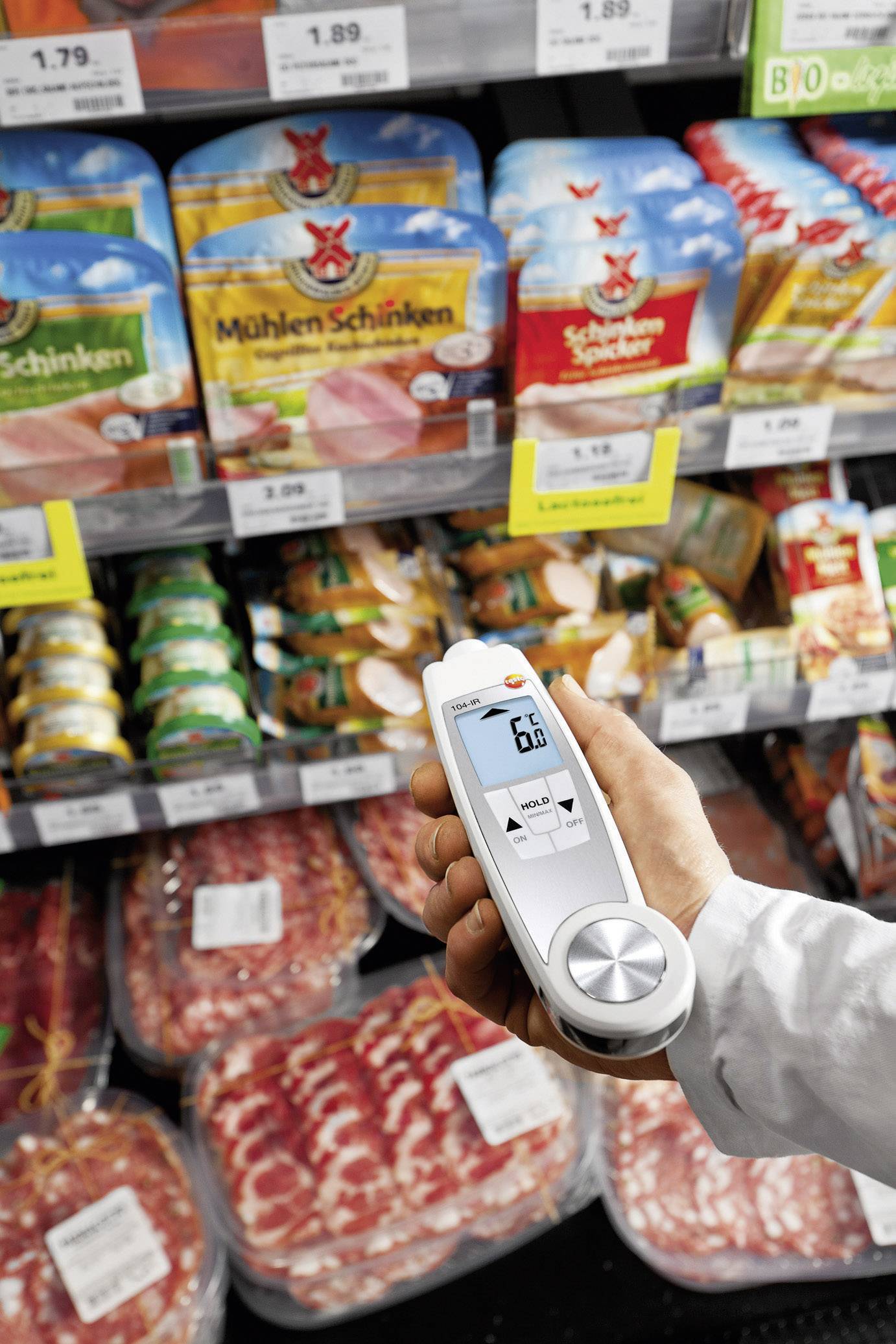 En person holder en digital temperaturmåler foran indpakket kød i køleskabet i et supermarked.