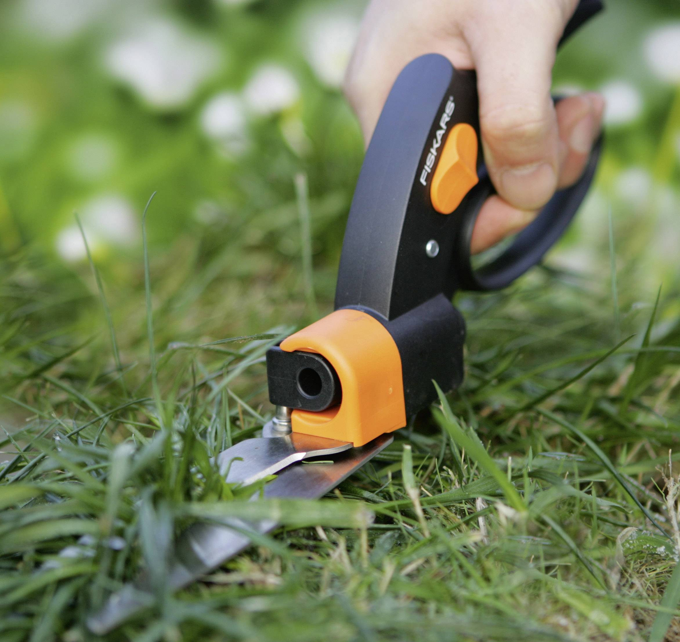 Hånd holder en orange-sort hækkeklipper og klipper præcist græsset i haven. Plænekatten passes, og scenen fremstår typisk sommerhyggelig udendørs.