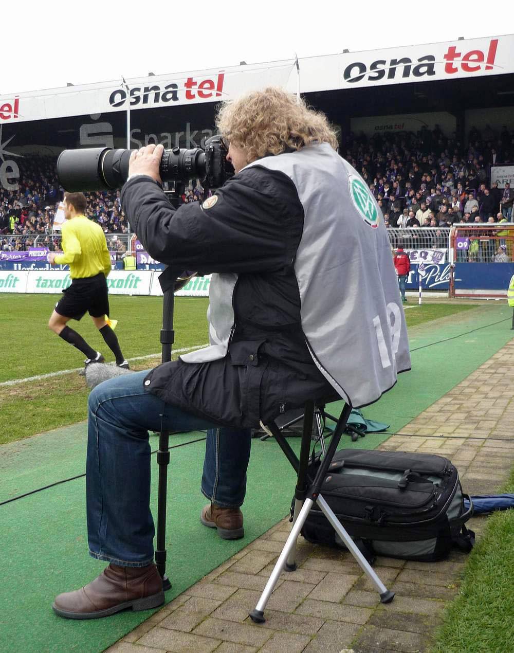 En fotograf med kamera og teleobjektiv sidder ved sidelinjen af en fodboldbane, mens tilskuere fylder tribunerne i baggrunden.