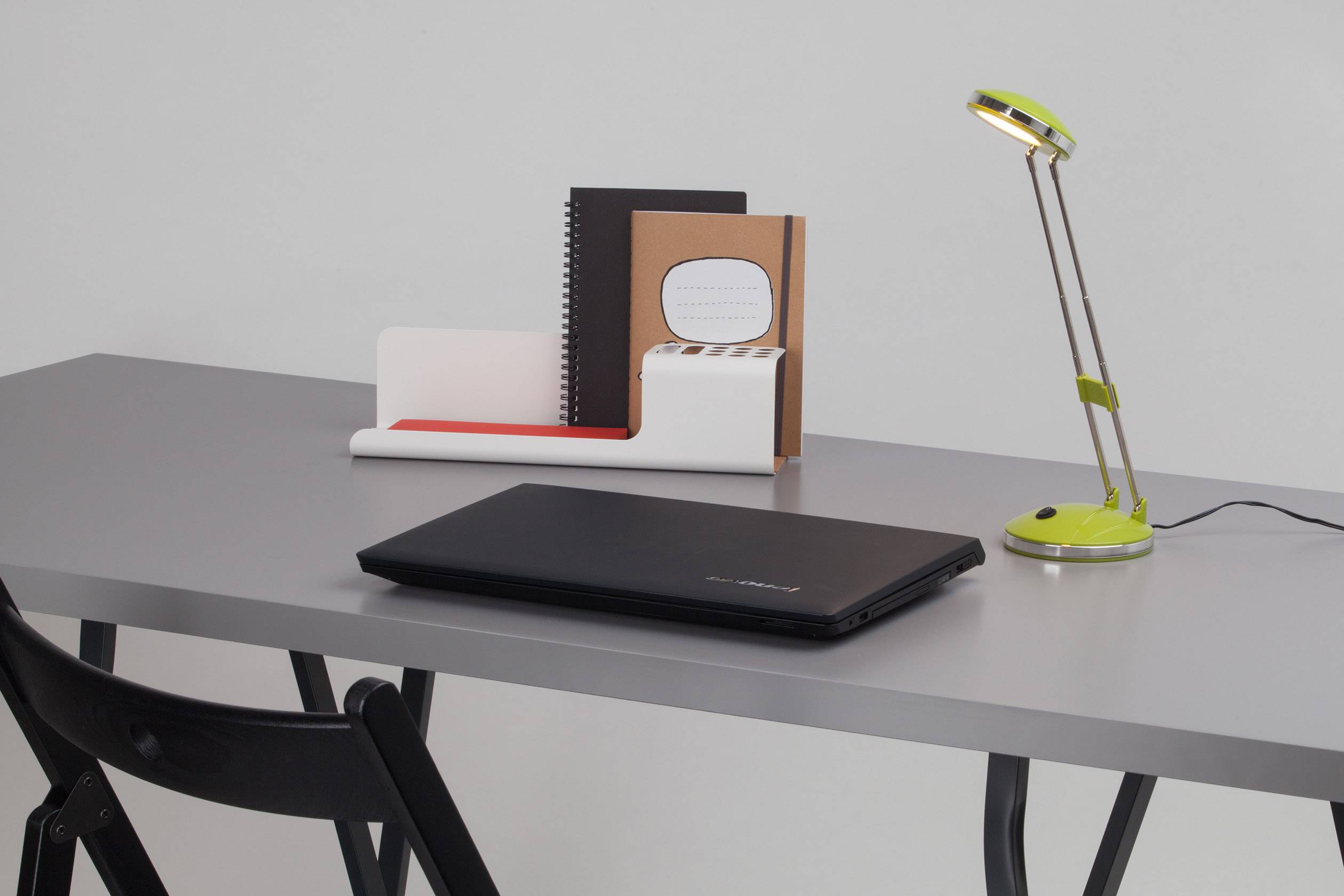 Et grå skrivebord med en sort bærbar computer, en bordlampe og en holder med mapper og notesbøger.
