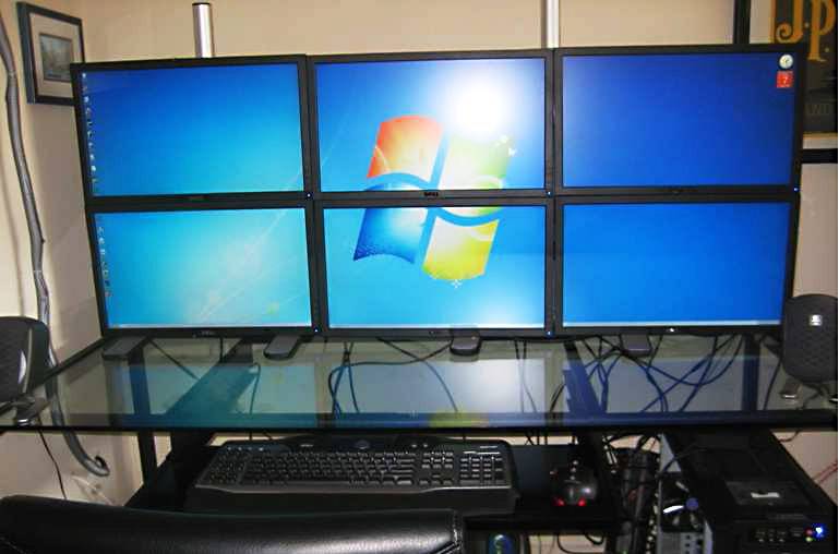 En arbejdsplads med seks skærme viser Windows-logoet på en blå baggrund. Et tastatur og en mus er placeret på skrivebordet.