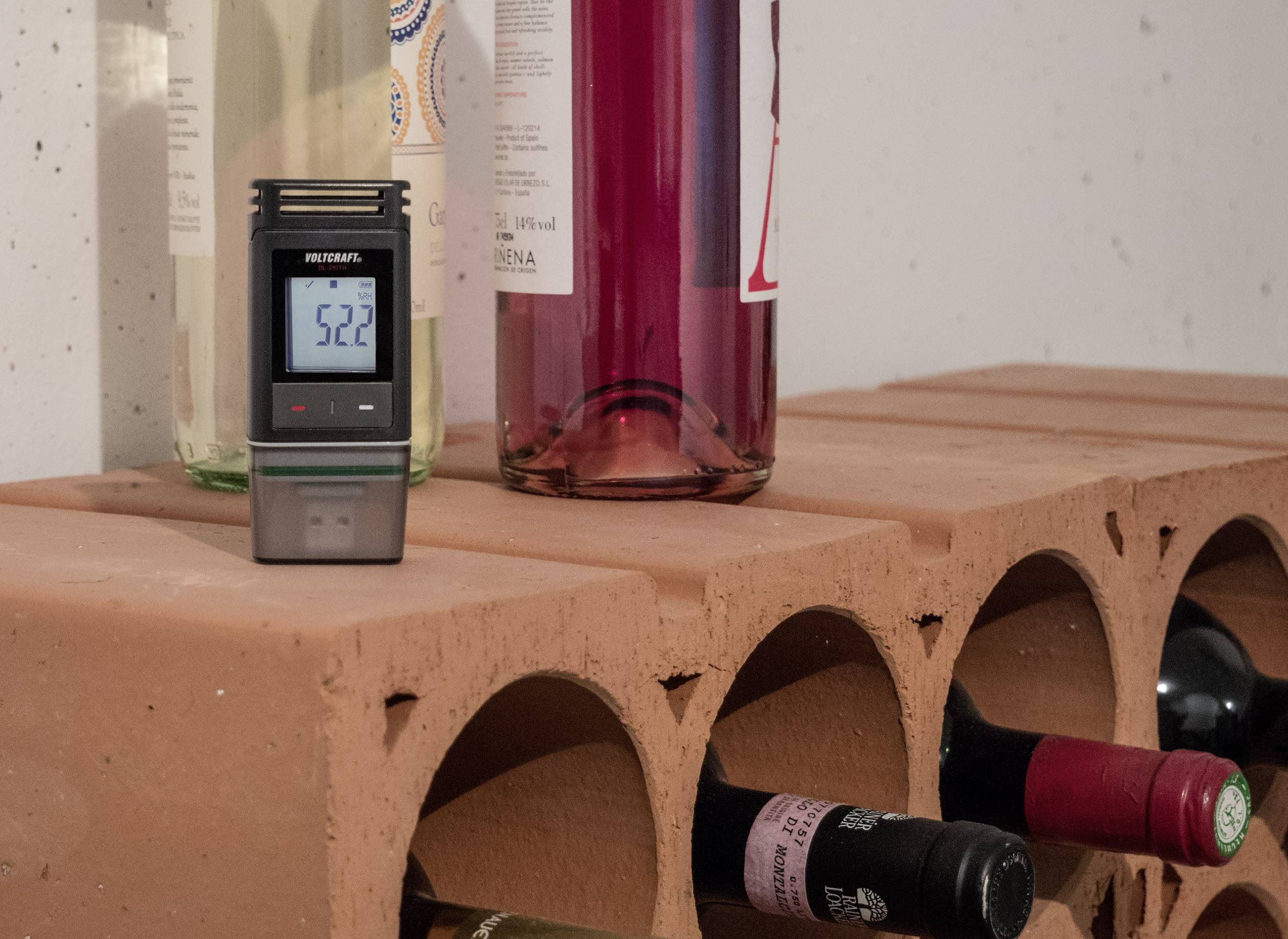 Et hygrometer viser 52,2 % luftfugtighed og står på en muret hylde med flere vinflasker.