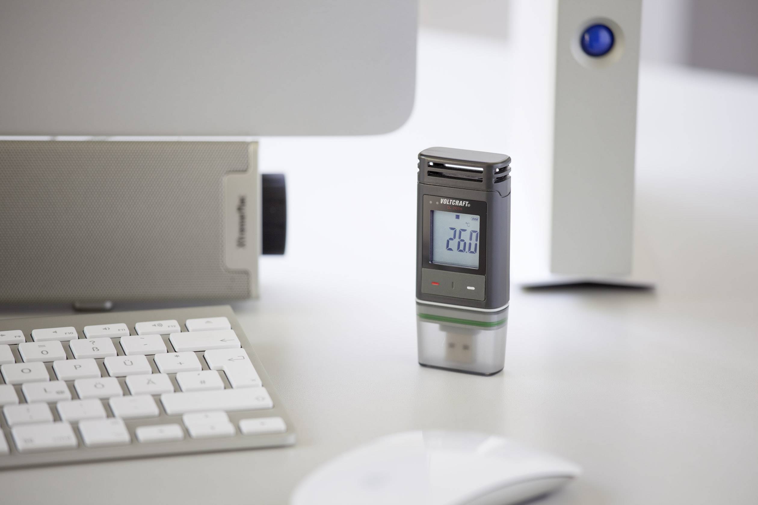 Et termometer med digitalt display viser '26°C'. Det står på et skrivebord tæt på en computerskærm og et tastatur.