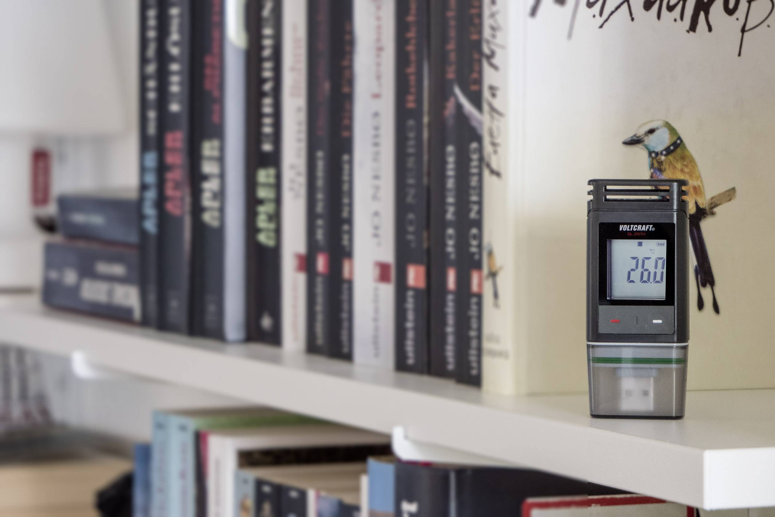 Et digitalt hygrometer viser '26.0' på en bogreol, omgivet af forskellige bøger.