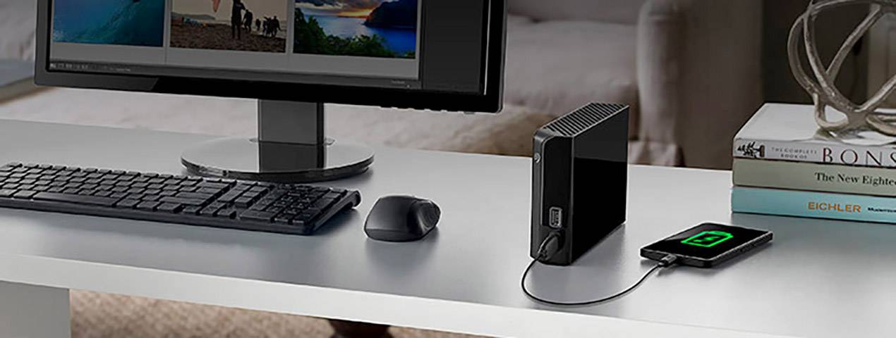 En computerskærm, et tastatur og en mus på et skrivebord. En smartphone forbindes via et kabel til en ekstern harddisk.