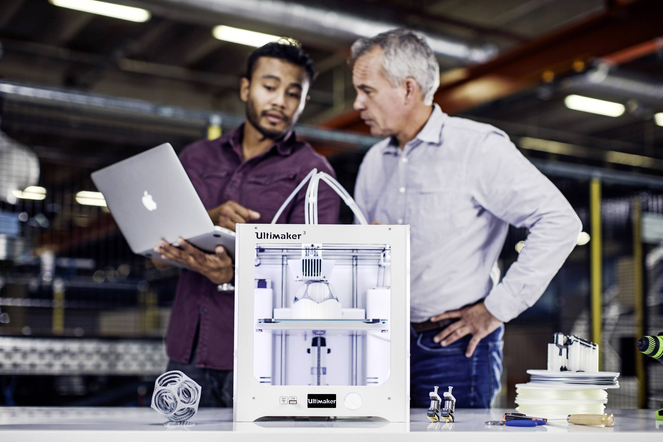 To mænd betragter en 3D-printer på et bord. Den ene holder en bærbar computer, og begge ser ud til at arbejde i et fabriksmiljø.