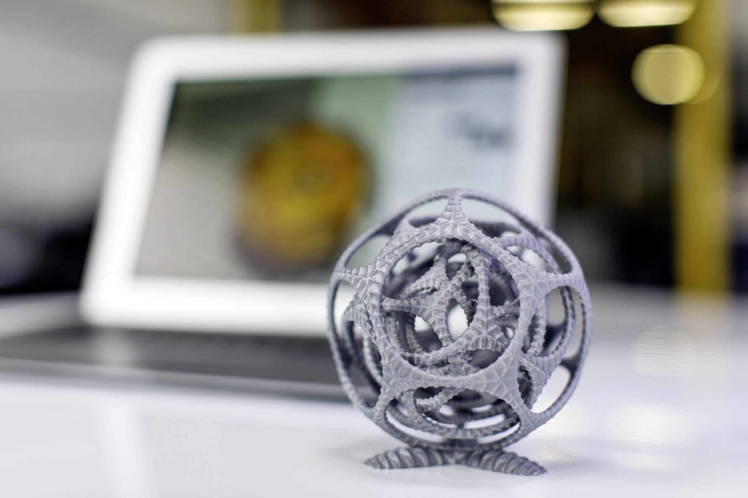 Grå, kompleks 3D-printet kugleobjekt i forgrunden; sløret bærbar computer i baggrunden viser et uskarpt billede.