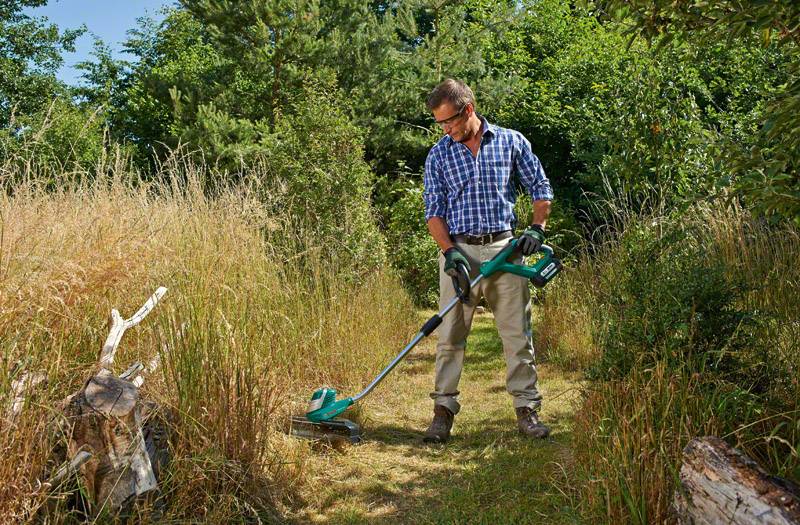 En mand i ternet skjorte og beskyttelsesbriller klipper græs med en trådløs græstrimmer i en overgrown have.